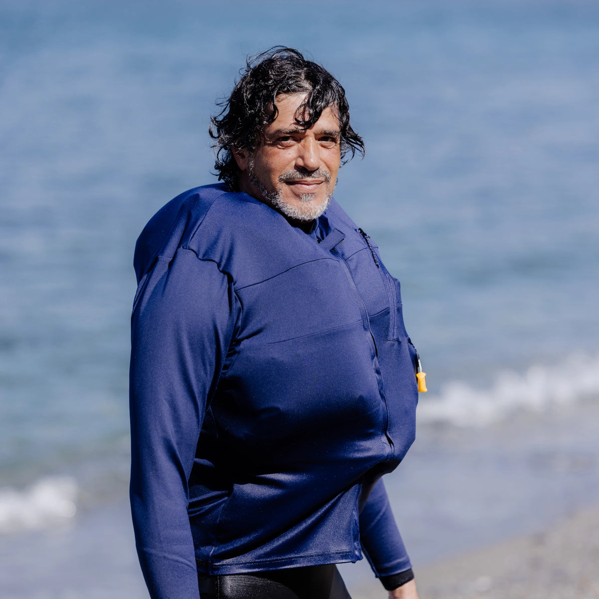 Man on a beach wearing a Floatee anti-drowning T-shirt for adults, color blue with long sleeves, with the inner life jacket inflated